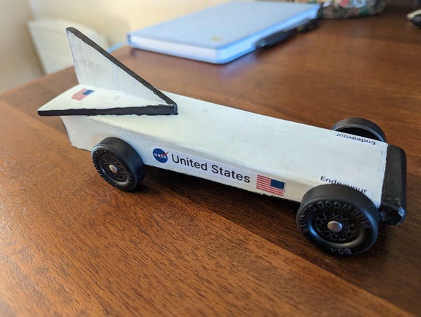 A pinewood derby car shaped and painted like the space shuttle Endeavour, white with black edging and an American flag an NASA logo.